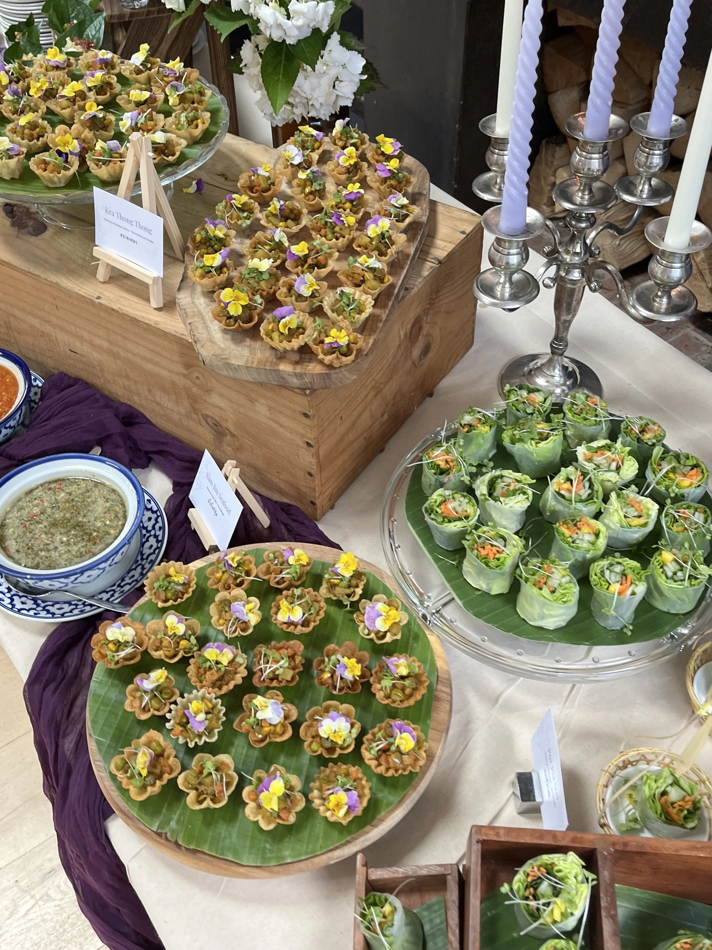 A display of various Thai appetizers, including small tartlets topped with edible flowers, fresh spring rolls filled with vegetables, and dipping sauces, arranged on colorful trays and a table with purple cloth. There is a silver candelabra with multicolored candles.
