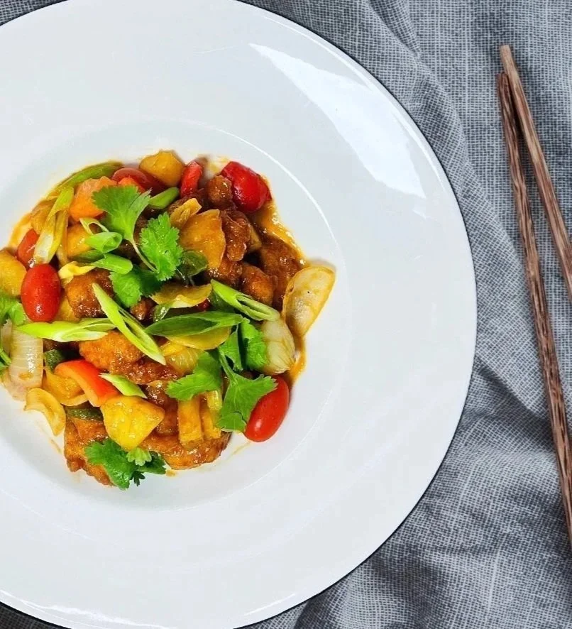 A white plate with a colorful stir-fried vegetable and meat dish, garnished with cilantro, on a gray fabric tablecloth with chopsticks placed beside the plate.