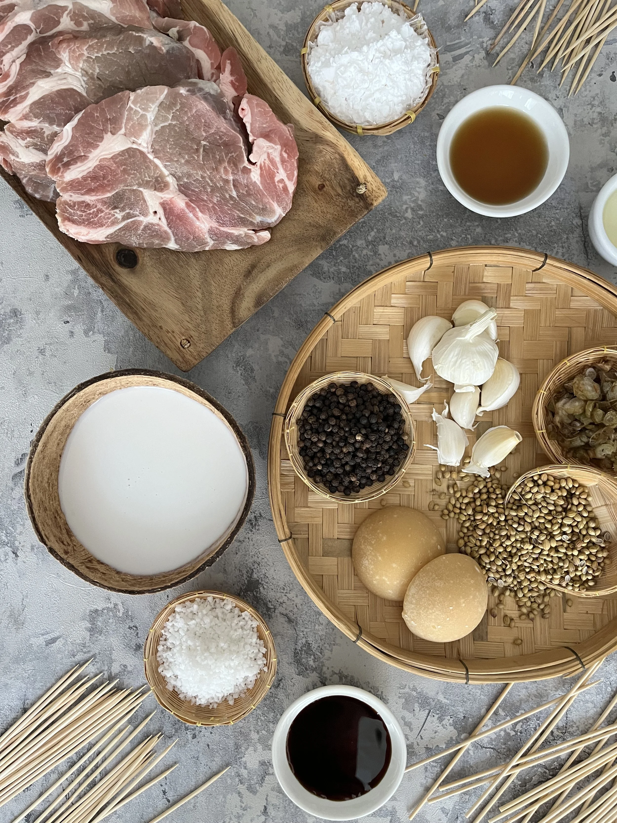 Ingredients for making skewers, including pork slices, garlic, black pepper, sliced coconut, and various sauces on a gray surface.