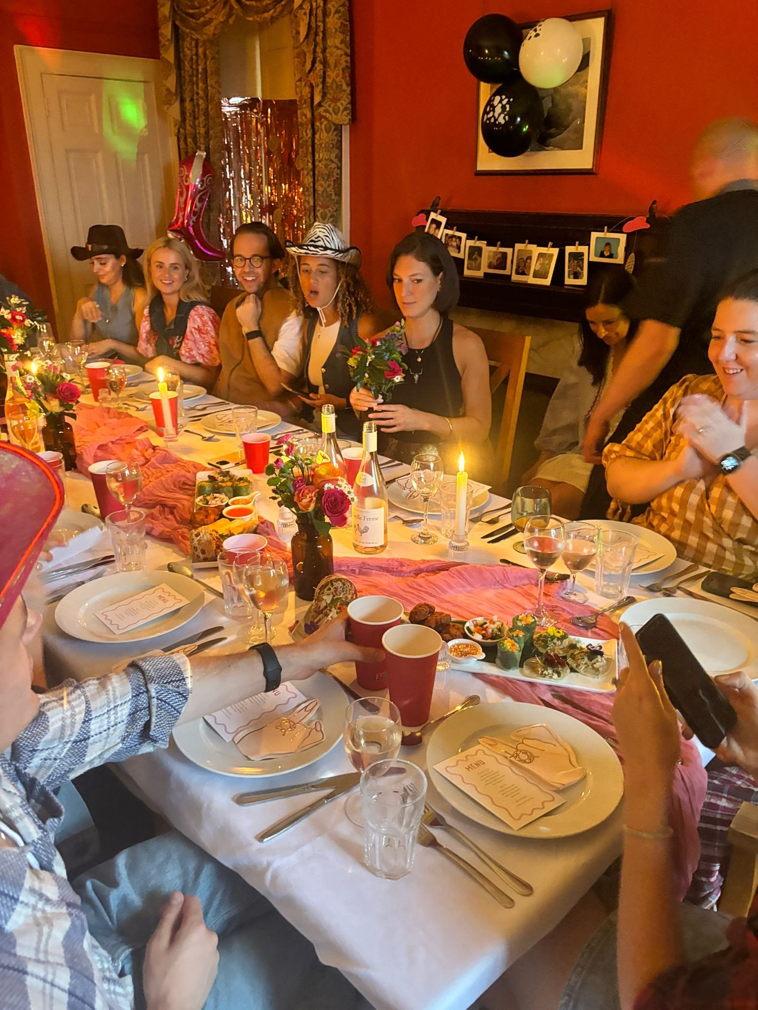 People gathered around a dining table celebrating with food, drinks, and decorations, including balloons and photos hanging on a string against a red wall.