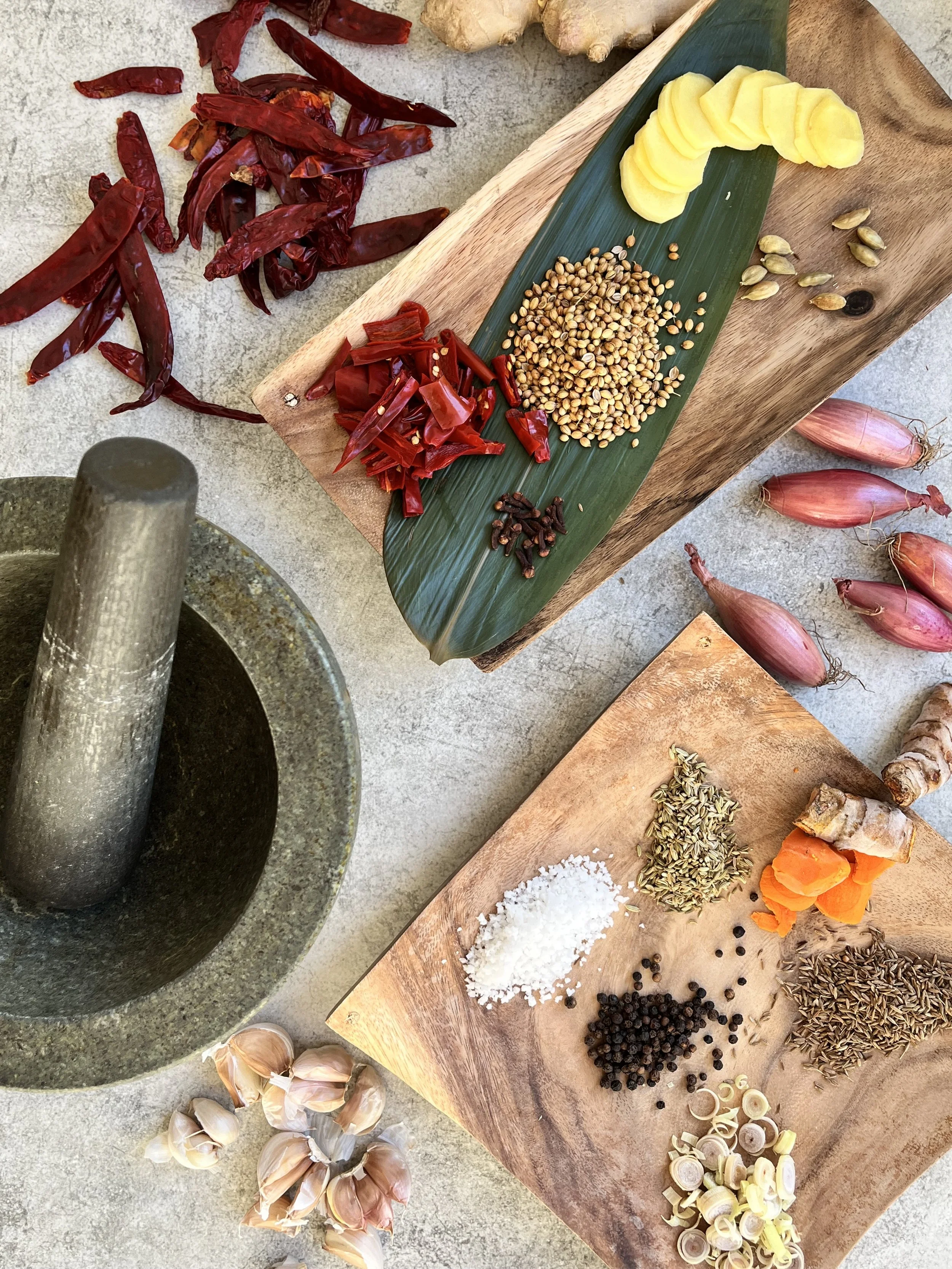 Various spices and ingredients on wooden and stone surfaces, including dried red chili peppers, sliced ginger, garlic, lemongrass, turmeric, and peppercorns, arranged on cutting boards and a mortar and pestle.