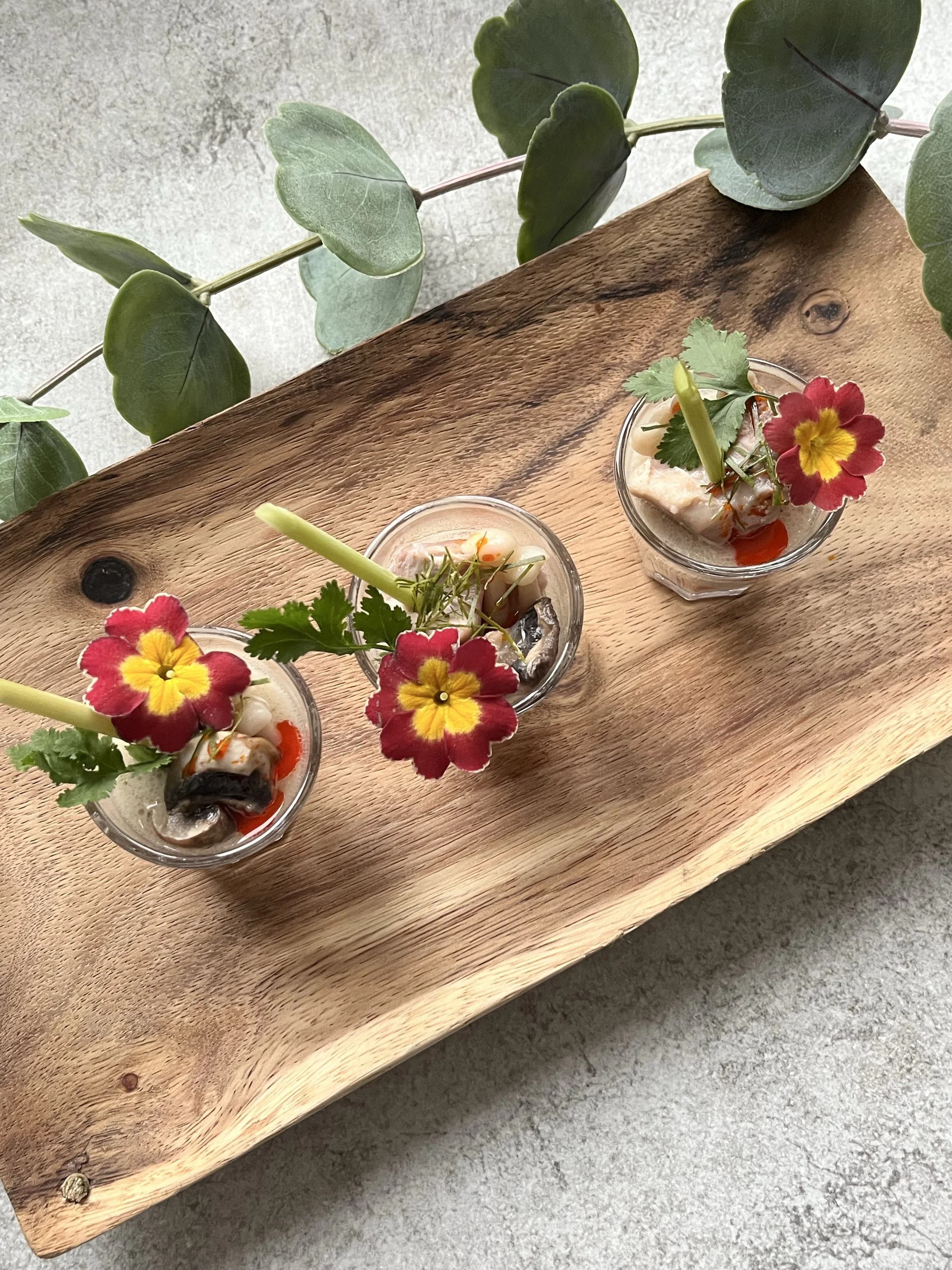Three small glasses of appetizer garnished with colorful edible flowers, green leaves, and celery sticks placed on a wooden tray.