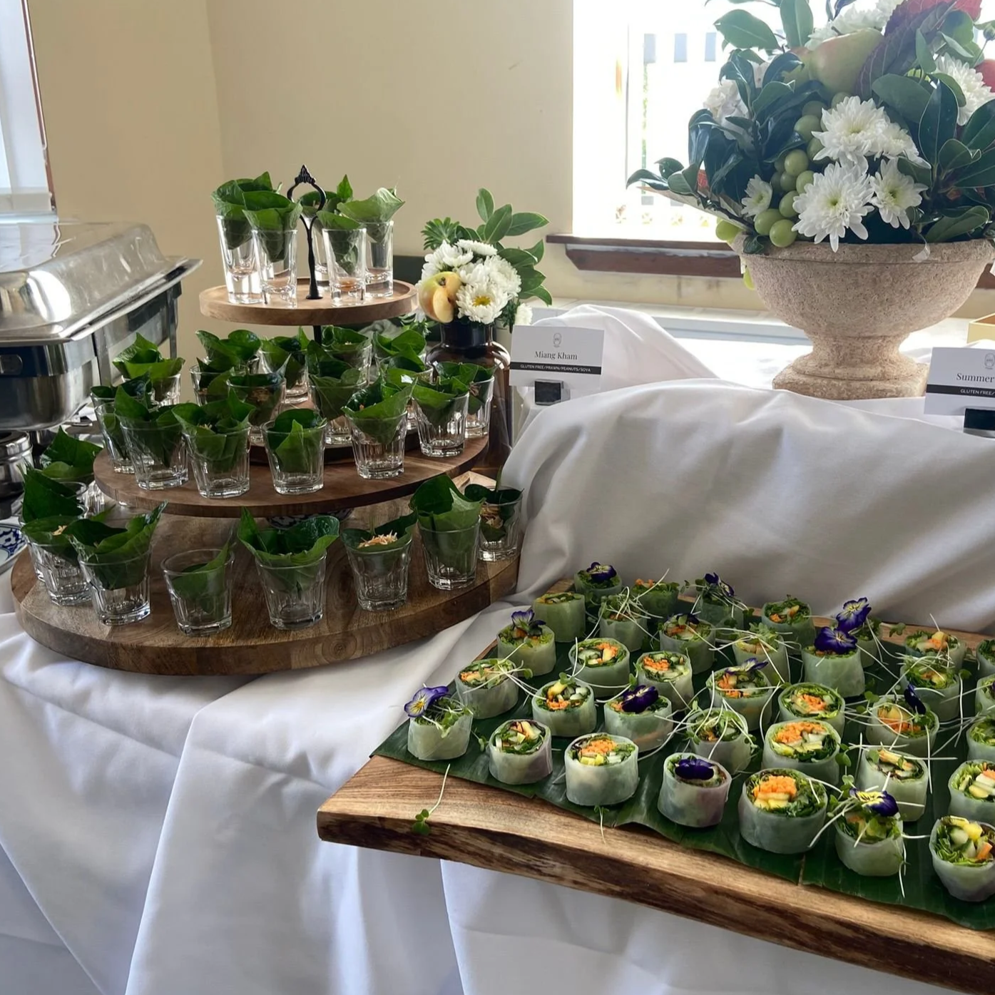 Display of appetizers and finger foods at a buffet, including vegetable spring rolls garnished with edible flowers, small glasses of green tea, and floral arrangements on a white tablecloth.