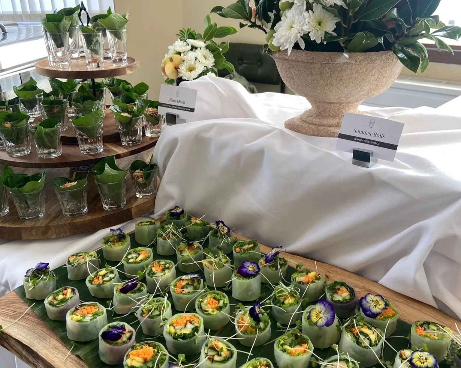 Assorted summer rolls garnished with edible flowers on a wooden tray, with glass cups and potted plants in the background at a buffet table.