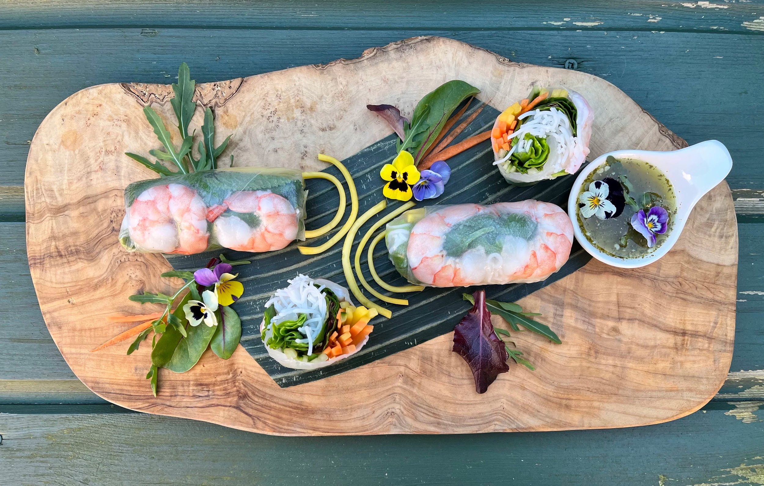 Fresh spring rolls with shrimp on a wooden platter, garnished with edible flowers, sliced vegetables, herbs, and dipping sauce.