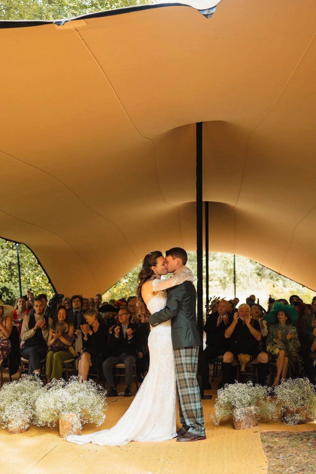a newlywed couple sharing a kiss under a wedding tent, with the groom wearing  a kilt and guests seated behind them.