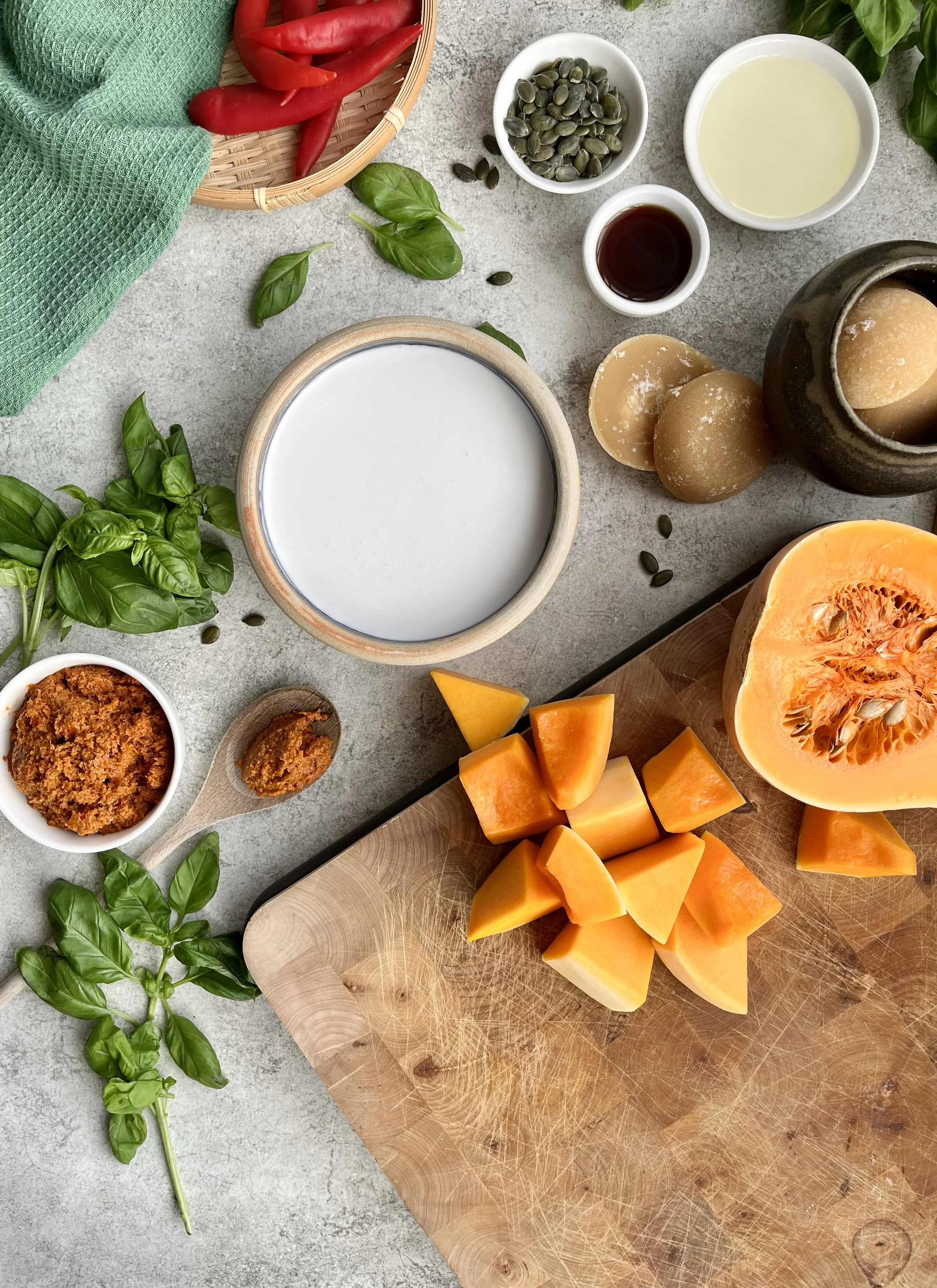 A variety of ingredients for a cooking or salad preparation, including chopped butternut squash, fresh basil, pumpkin seeds, chili peppers, a jar of brown sugar, a bowl of creamy white liquid, a small bowl of dark sauce, a bowl of pumpkin seeds, and a piece of butternut squash with seeds, all arranged on a gray surface.