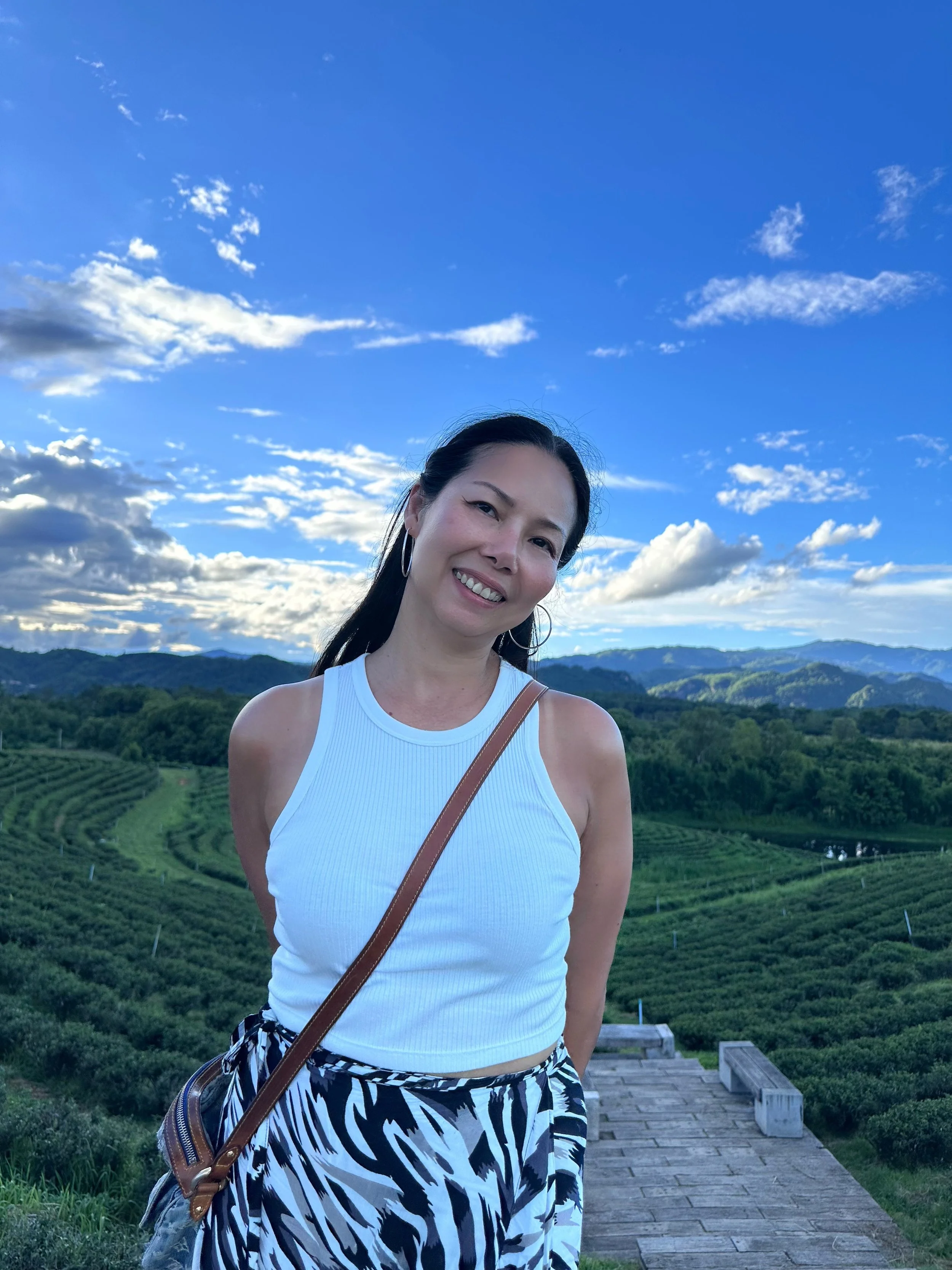 Smiling woman standing outdoors with rolling hills and a bright blue sky with clouds in the background.