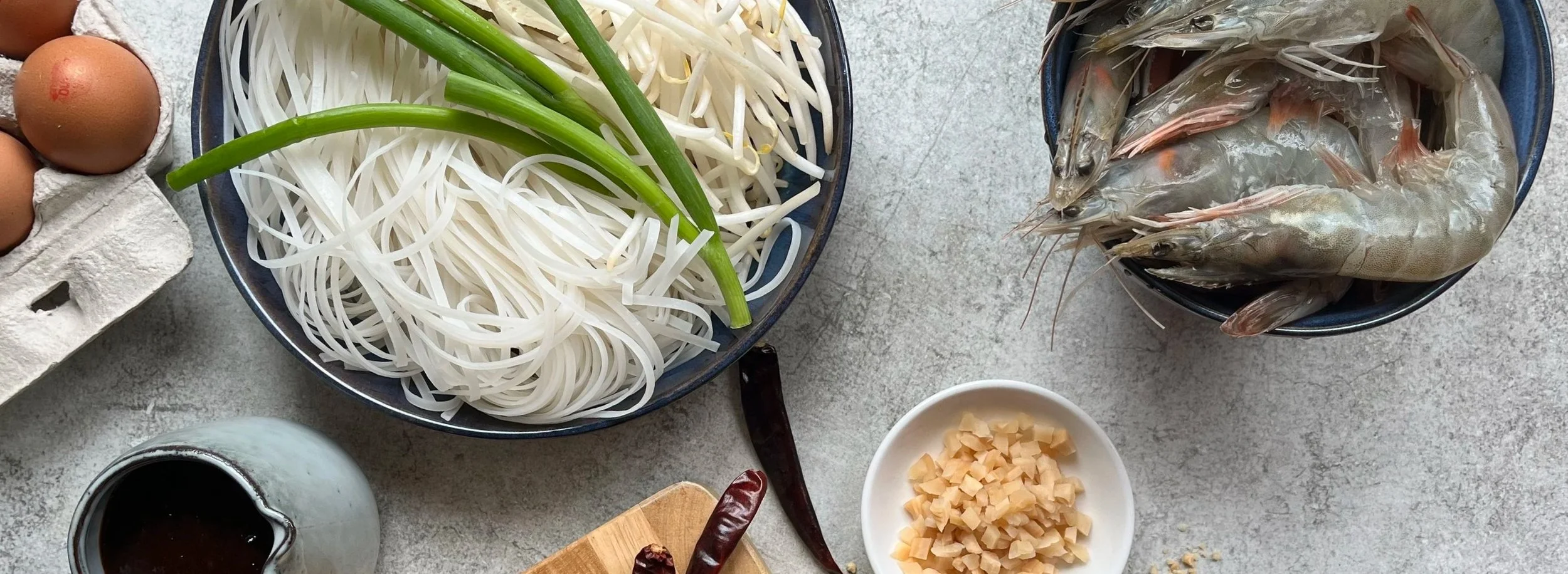 Layout of ingredients including eggs, rice noodles, bean sprouts, green onions, shrimp, chopped garlic, soy sauce, and dried red chili peppers on a grey surface.