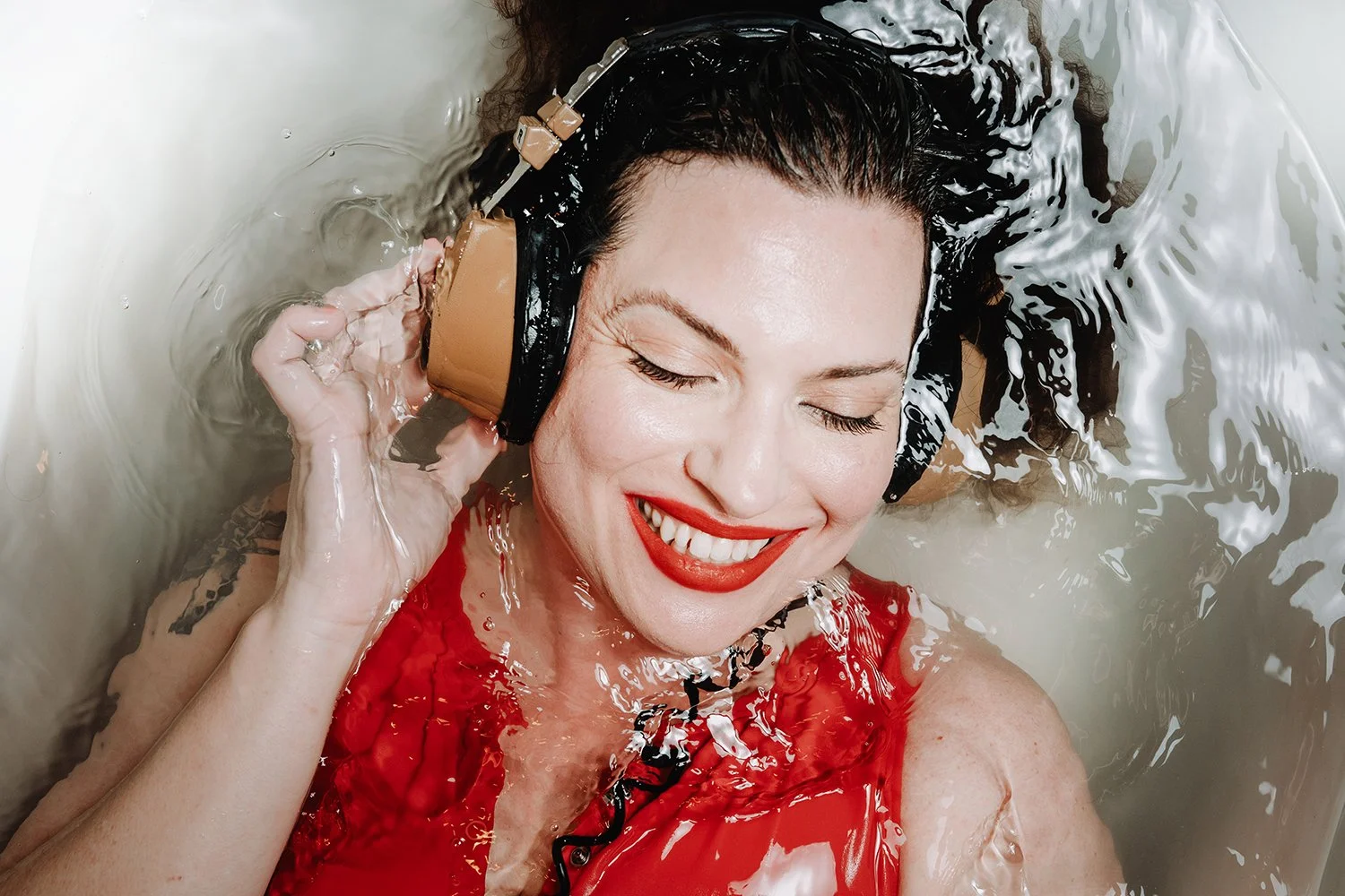 A woman with bright red lipstick and a red dress is smiling with eyes closed, wearing vintage-style headphones in water.