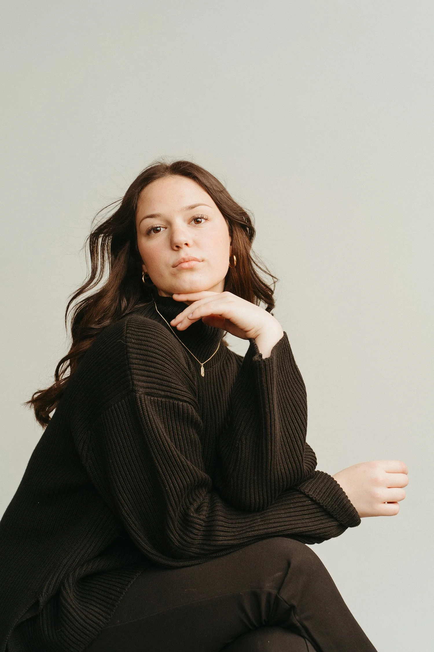 A young woman with wavy brown hair, wearing a black turtleneck sweater, golden hoop earrings, and a delicate necklace, sits with her hand resting under her chin against a plain light-colored background.