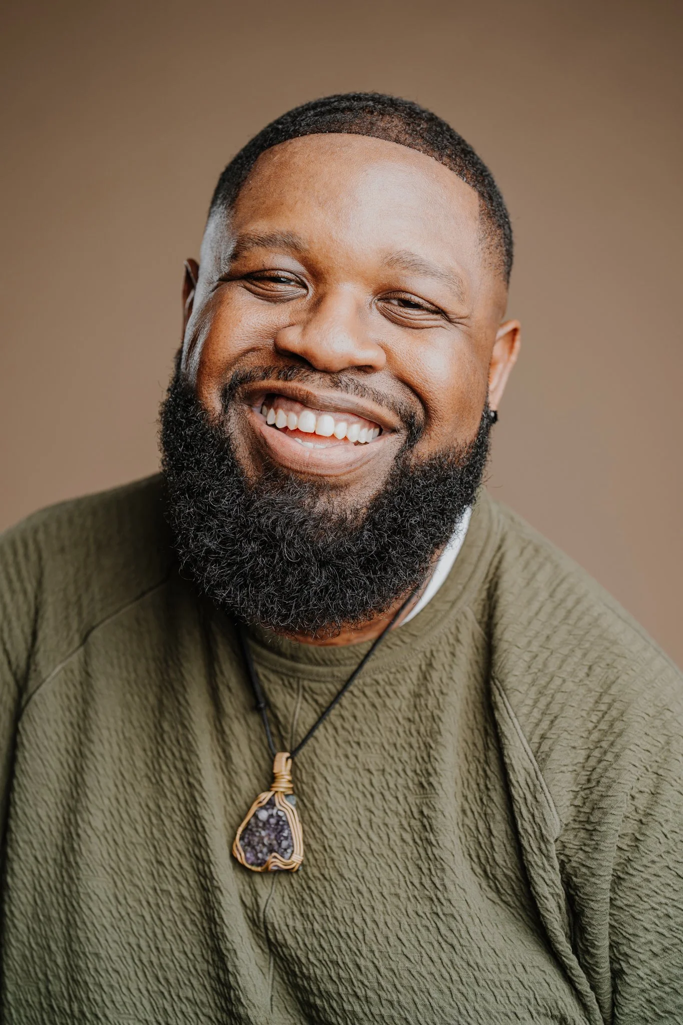 Close-up of a smiling African American man with a beard, wearing a green textured shirt and a pendant necklace with a purple stone.
