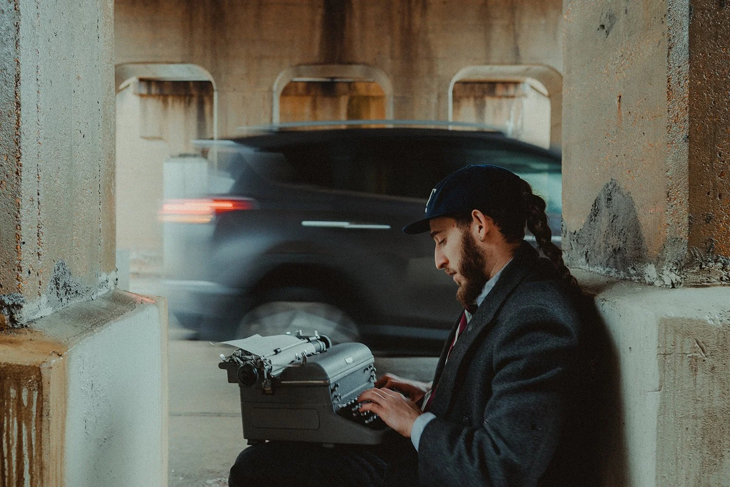 A man with a beard, wearing a dark coat, a plaid shirt, and a backward baseball cap, is sitting under a bridge, typing on a vintage typewriter. A moving black car is passing by behind him.