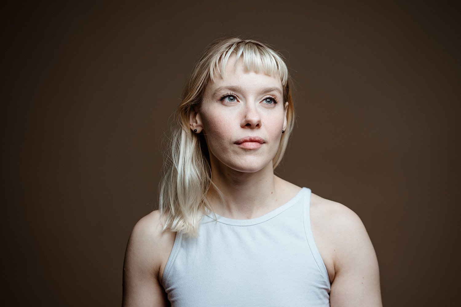 Portrait of a young woman with blonde hair and blue eyes looking slightly to the right, wearing a white tank top, against a brown background.