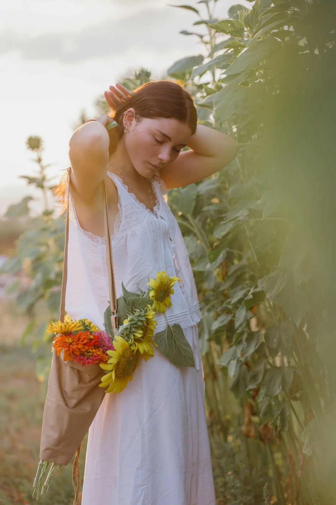 A young woman with brown hair in a white dress, standing in a sunflower field during sunset, with her eyes closed and hands on her neck, carrying a shoulder bag decorated with sunflowers and colorful flowers.