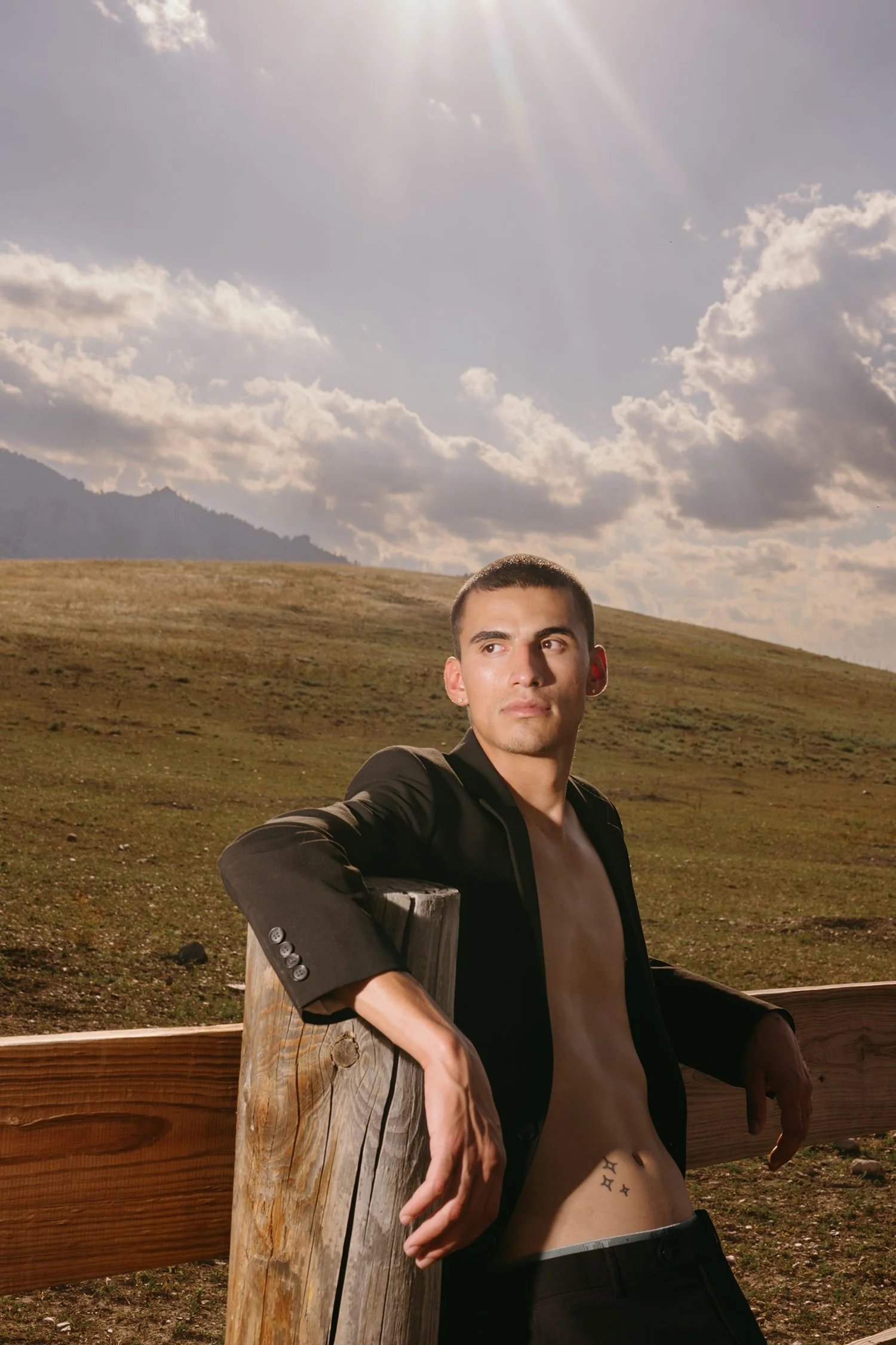 A young man leaning against a wooden fence with a landscape of rolling grassy hills and mountains in the background, under a partly cloudy sky with sunlight rays.