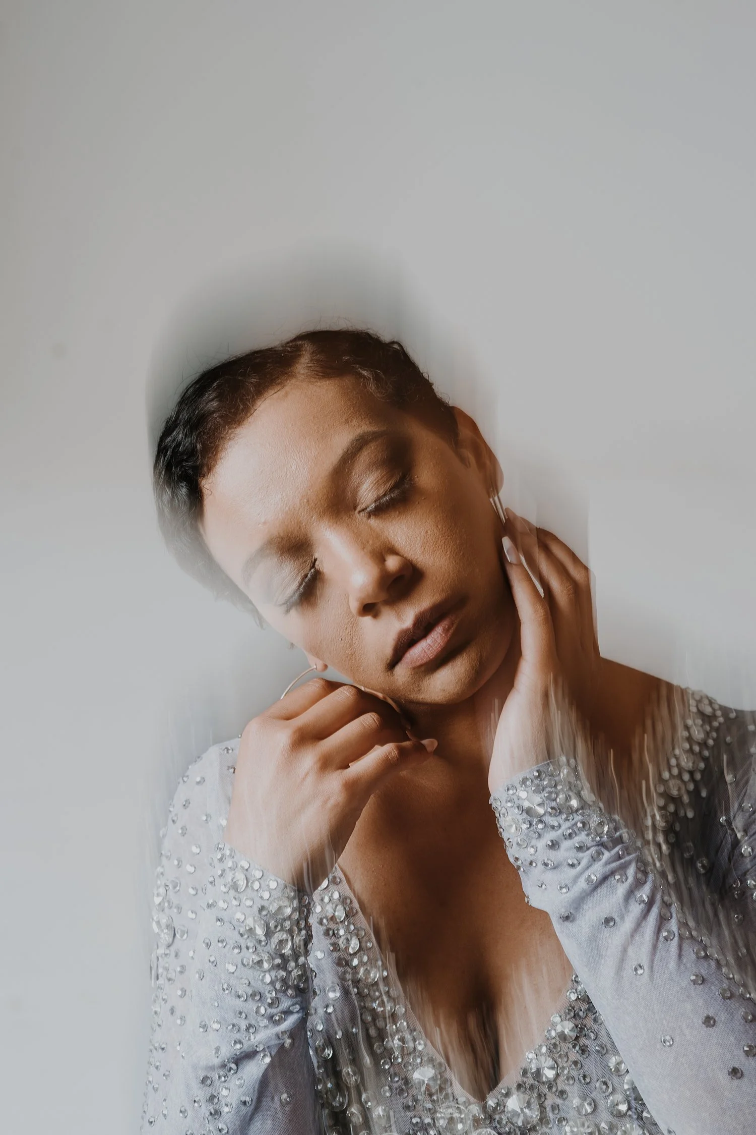 A woman with short dark hair, wearing a long-sleeved top decorated with pearls and rhinestones, has her eyes closed and touches her neck with both hands.