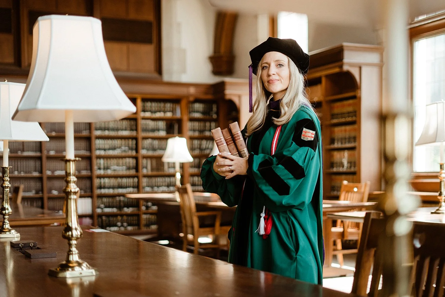 Woman in graduation gown and cap holding books inside a library with wooden bookshelves and tables.