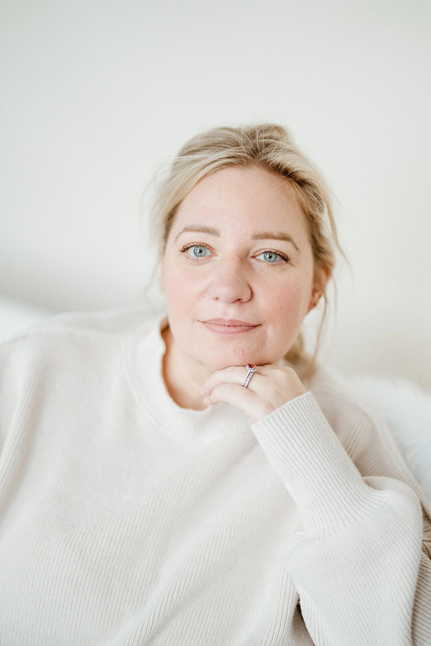 Close-up of a woman with blonde hair and blue eyes, wearing a white sweater, resting her chin on her hand, with a gentle smile, indoors against a white background.