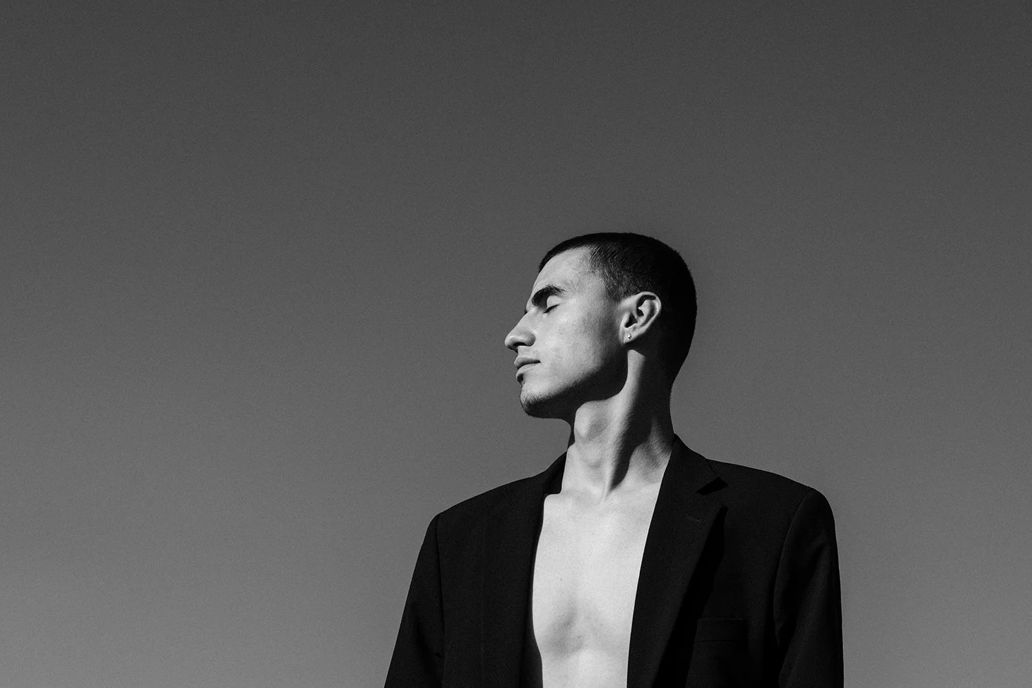 A black and white photo of a man with short hair and a goatee, facing sideways with eyes closed, wearing a partially open blazer with no shirt underneath, outdoors against a clear sky.