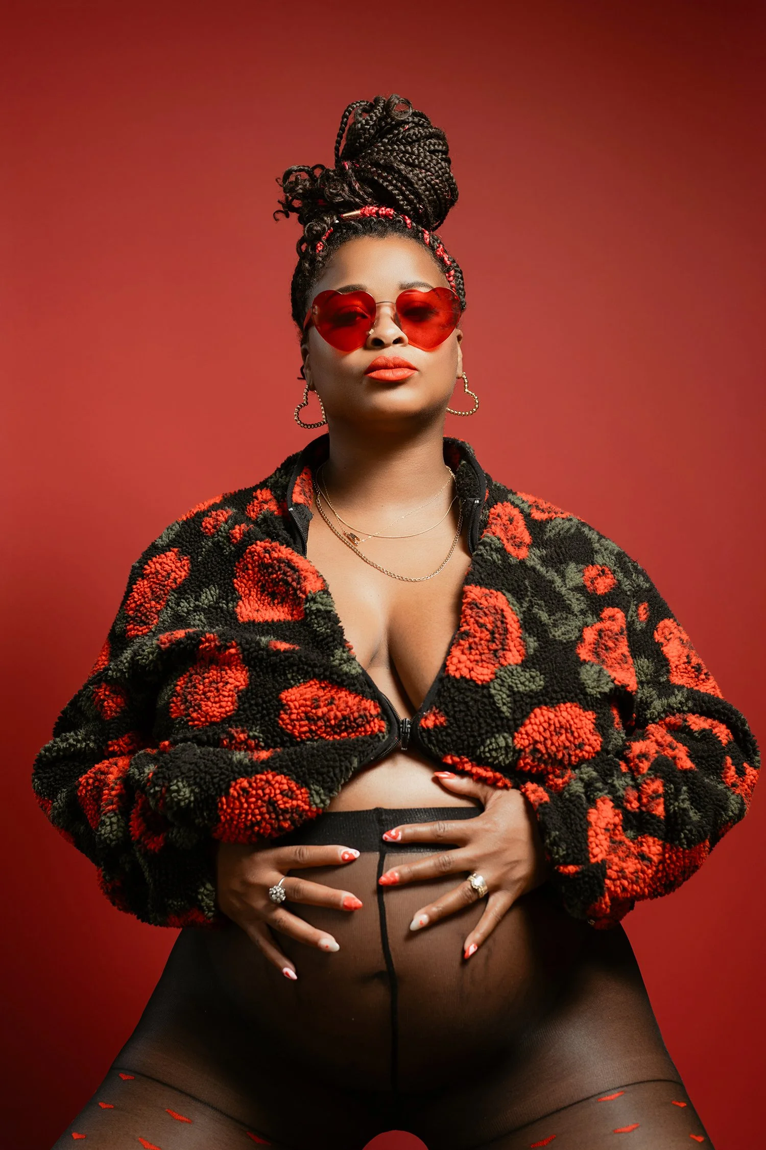 A woman with braided hair styled in a high bun, wearing red heart-shaped sunglasses, earrings, layered necklaces, a floral-patterned fleece jacket, sheer tights with red accents, and glossy makeup, posed against a red background.