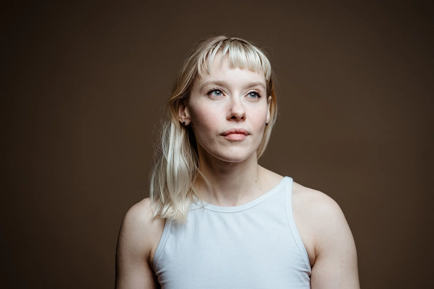 Portrait of a young woman with blonde hair wearing a white sleeveless top against a plain brown background.