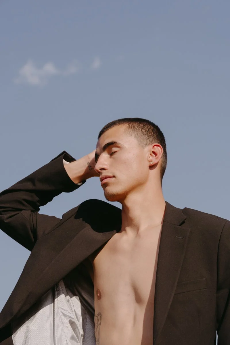 A young man with short hair, wearing a black blazer and no shirt underneath, standing outdoors against a blue sky with a few clouds, with his eyes closed and one hand behind his head.