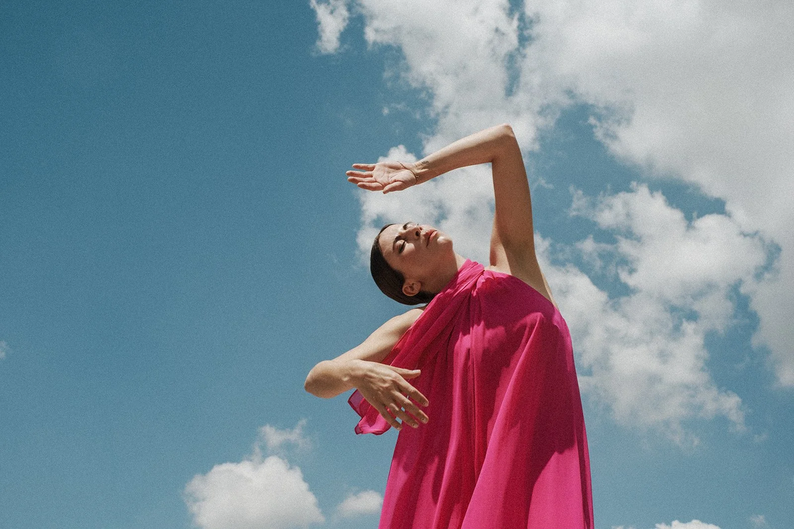 A woman in a pink dress standing outdoors against a blue sky with clouds, with her eyes closed and one arm raised above her head, striking a graceful pose.