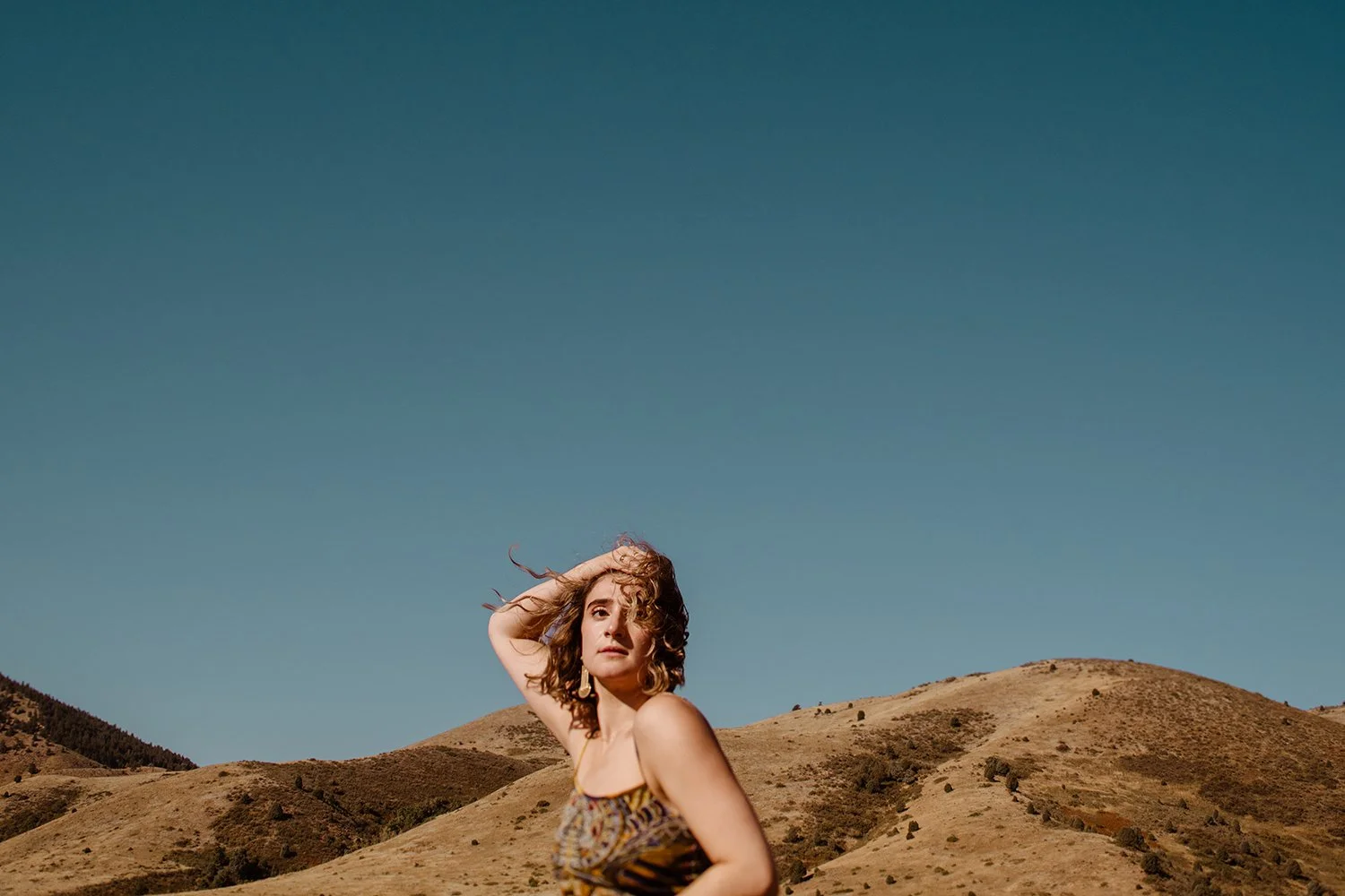 A woman with curly hair standing outdoors in a desert-like landscape with rolling hills and a clear blue sky.
