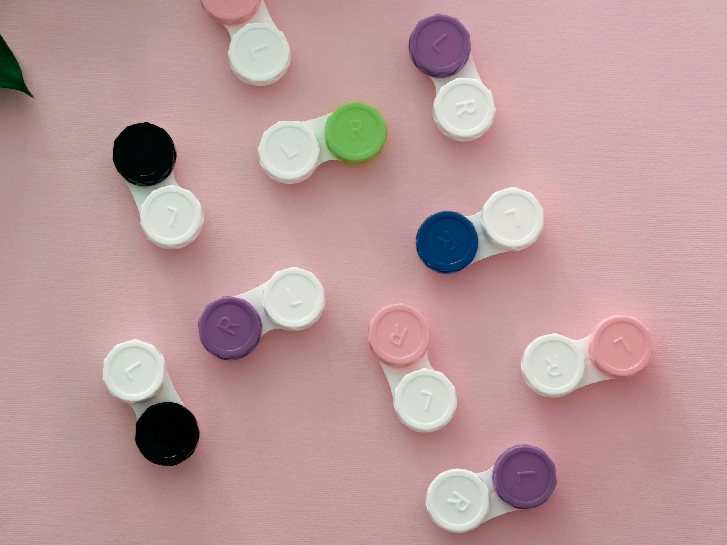 Multiple pairs of colored contact lens cases scattered on a pink surface, with each case labeled with 'L' or 'R' and a number or letter, in various colors including purple, blue, green, pink, white, and black.