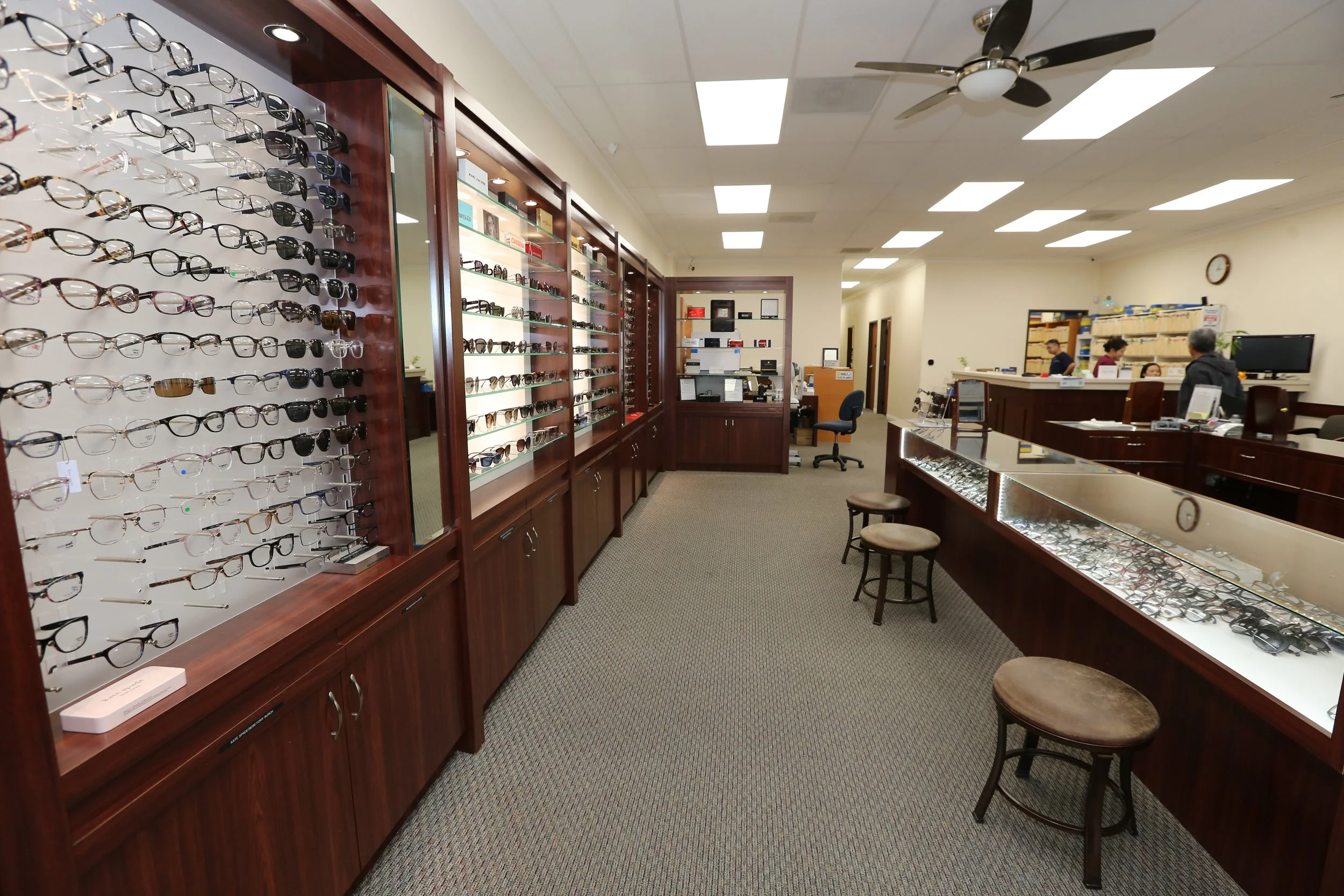 Interior view of an eyewear store with glasses displayed on wooden shelves and counters, a ceiling fan, and several customers and staff at the counter.