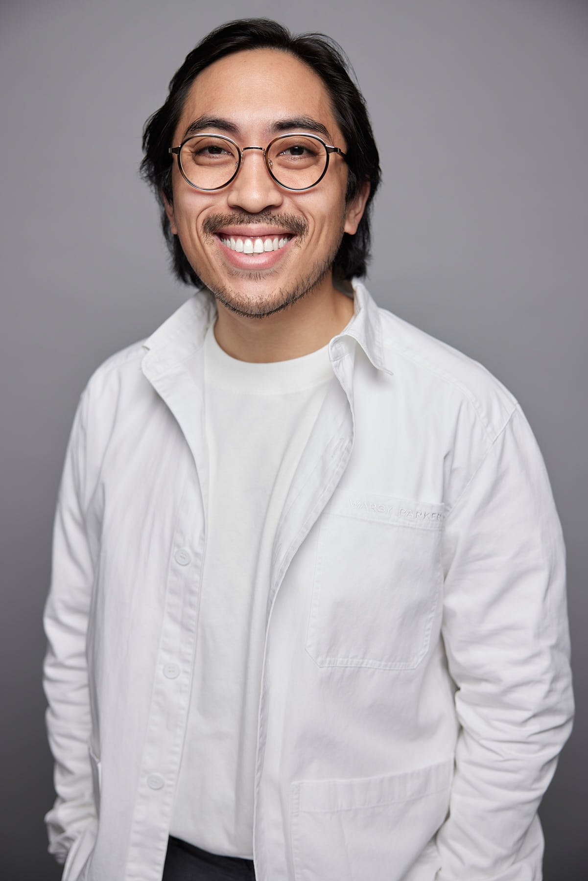 A smiling man with glasses and long dark hair against a gray background.