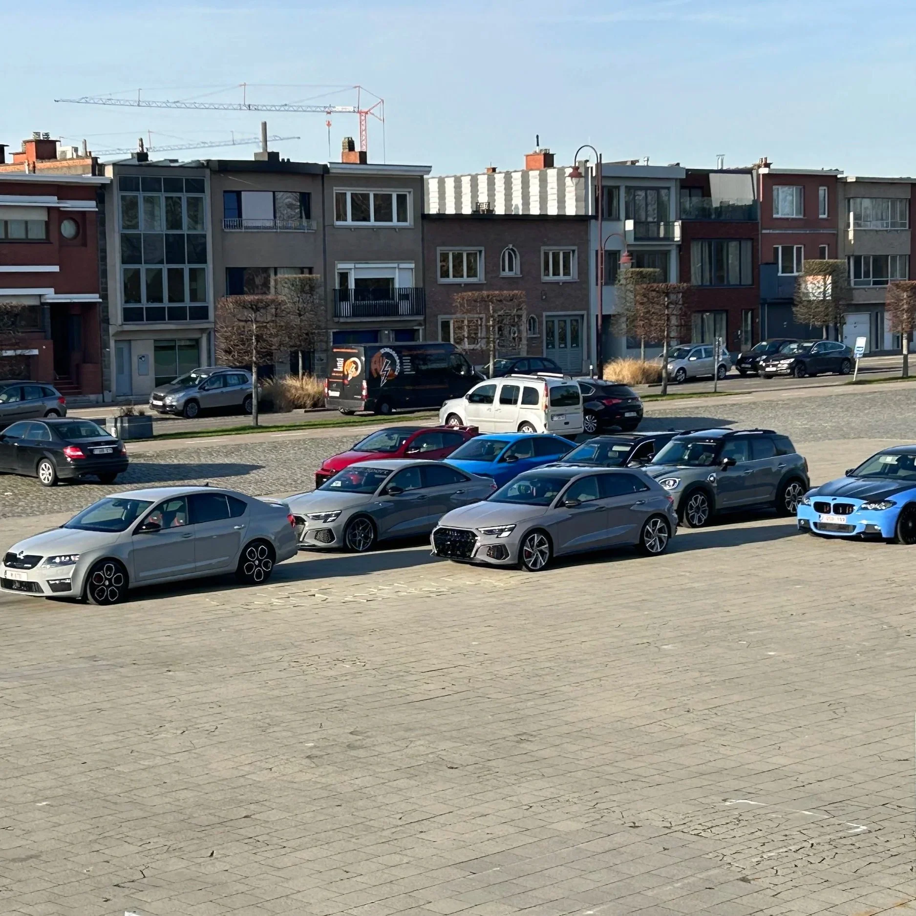 Multiple cars parked in an outdoor lot with modern apartment buildings and trees in the background.