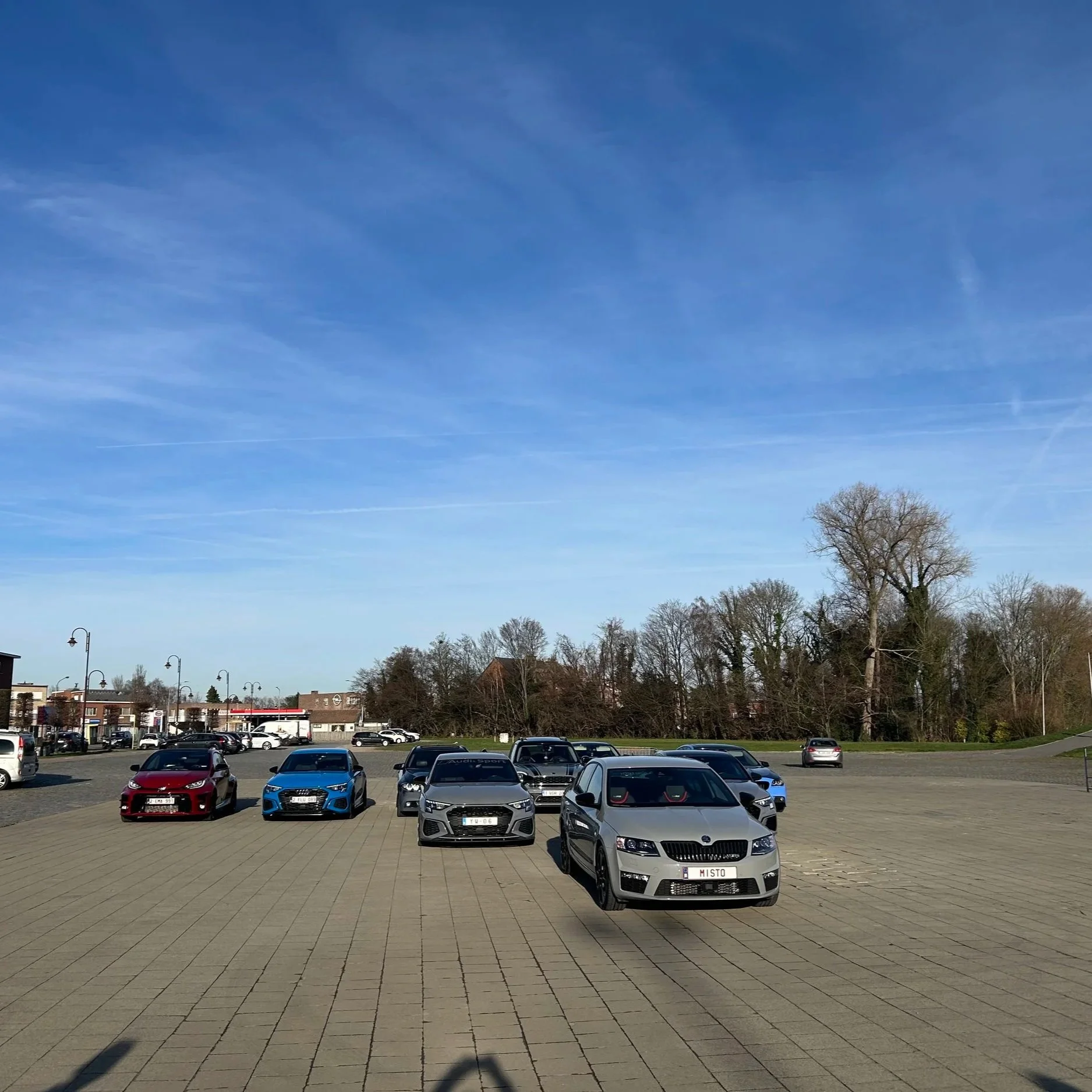 A parking lot with several cars, including a silver car in the front, under a clear blue sky with trees and buildings in the background.