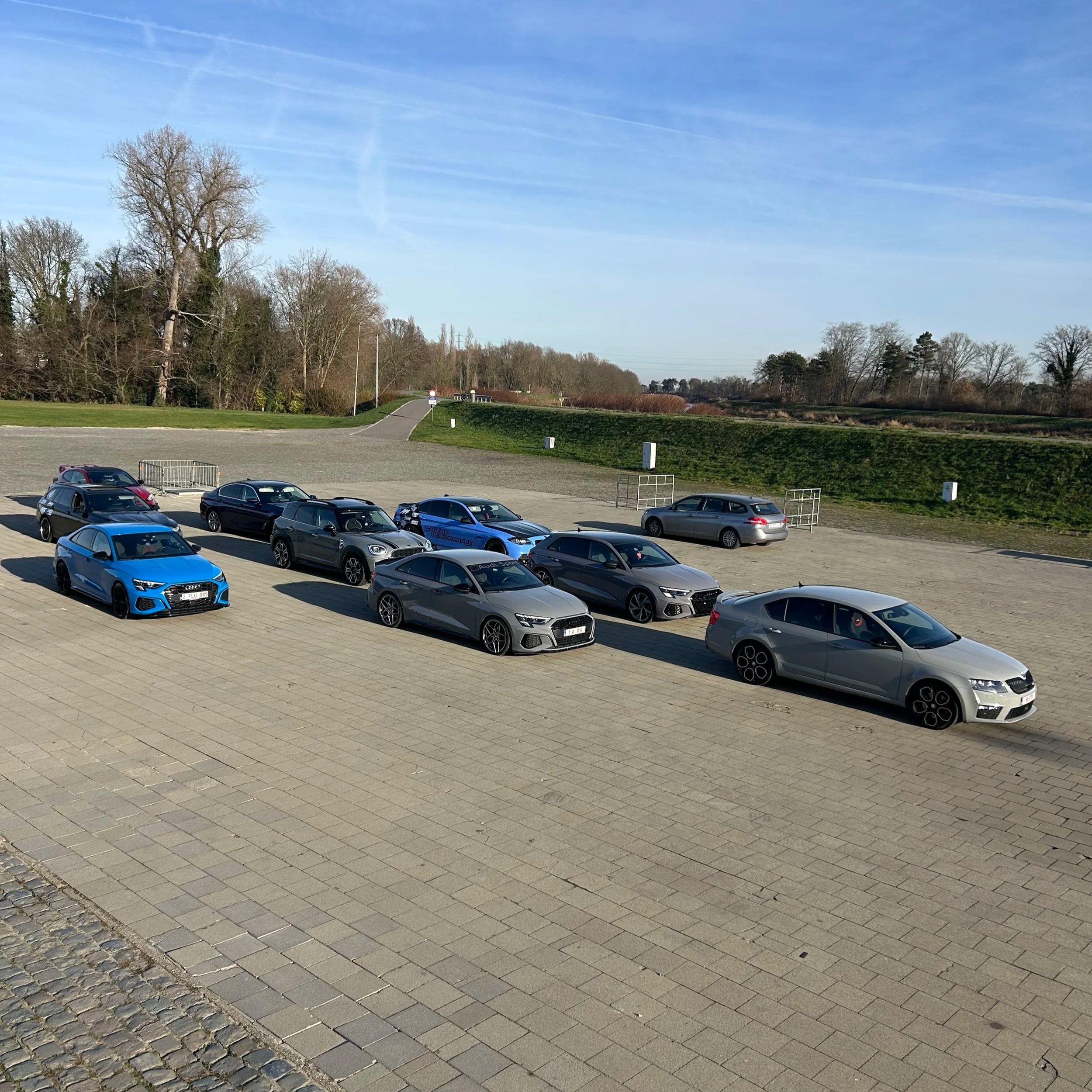 A parking lot with eleven parked cars of various colors, including blue, black, gray, and silver, under a clear blue sky with trees and a grassy hill in the background.