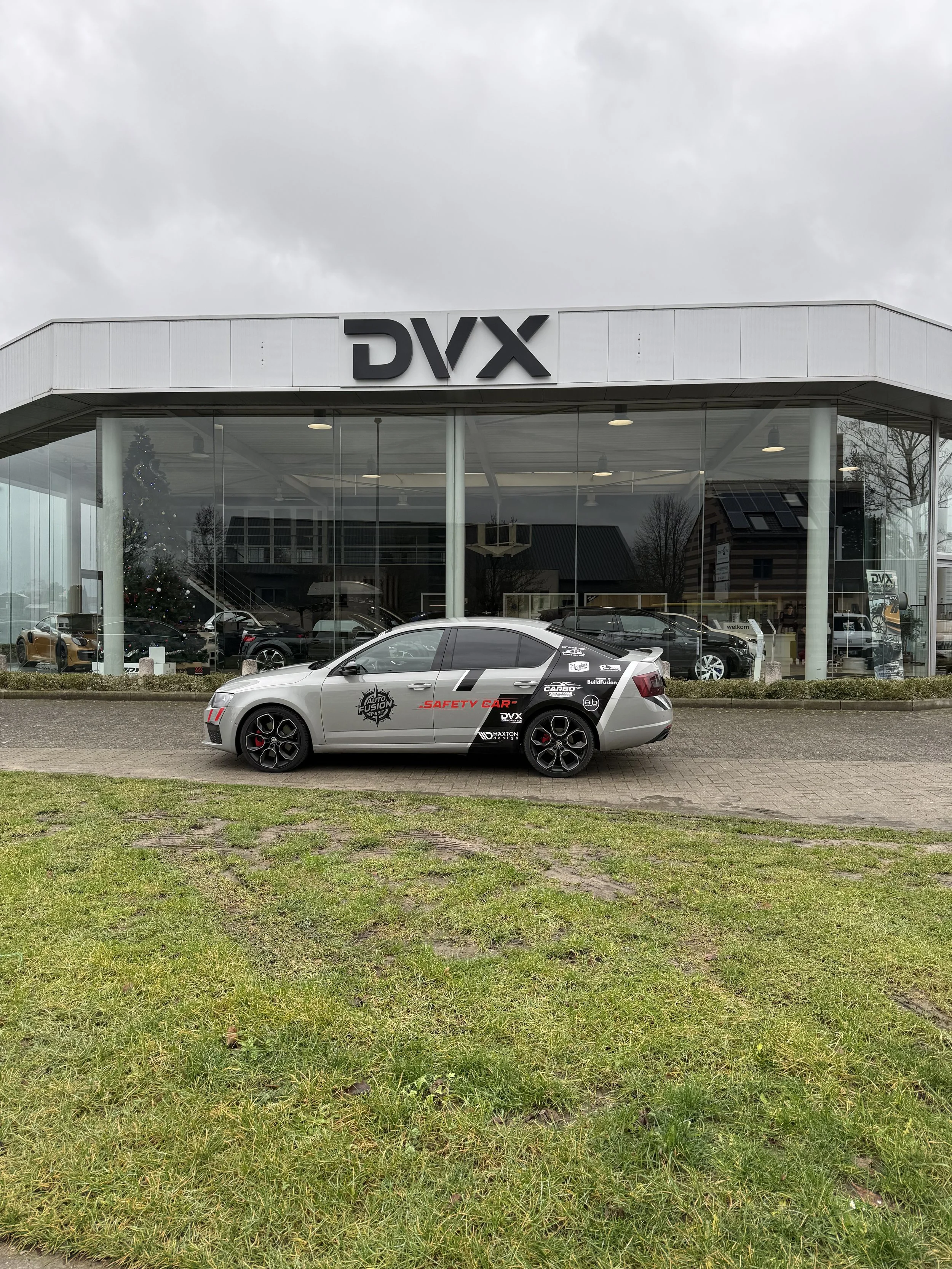 Car parked outside DVX dealership with a large glass showroom building behind it, displaying multiple cars inside. The car has branding related to safety testing and auto fusion, with various logos and text.