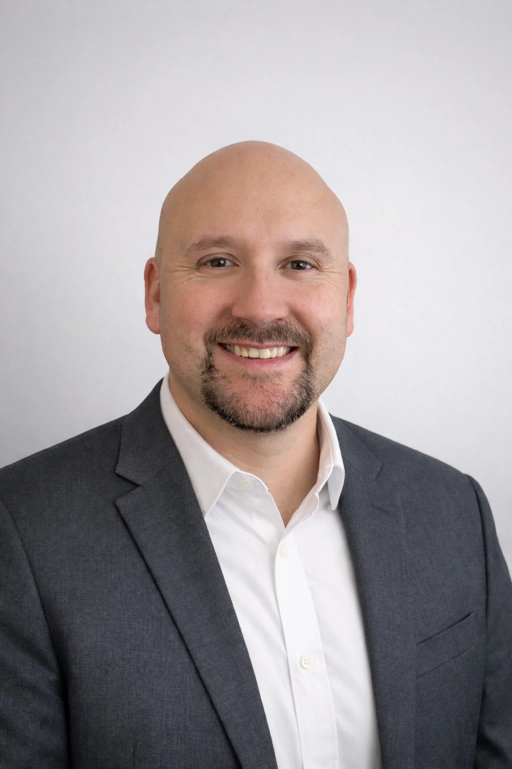 Headshot of a bald man with a goatee, wearing a dark suit and white shirt, smiling against a plain white background.