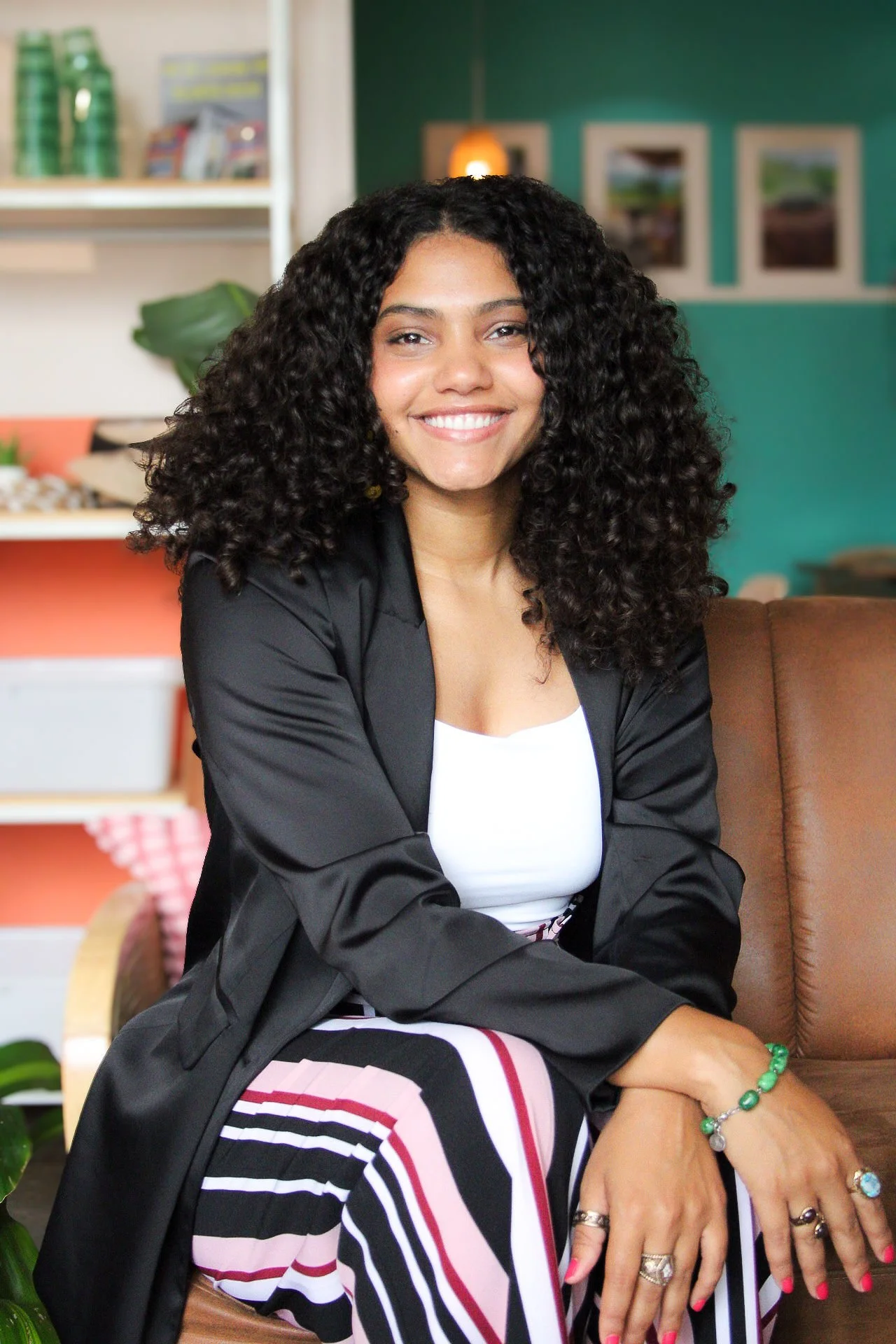 A woman with curly hair, smiling, sitting on a brown chair in a cozy, colorful room.