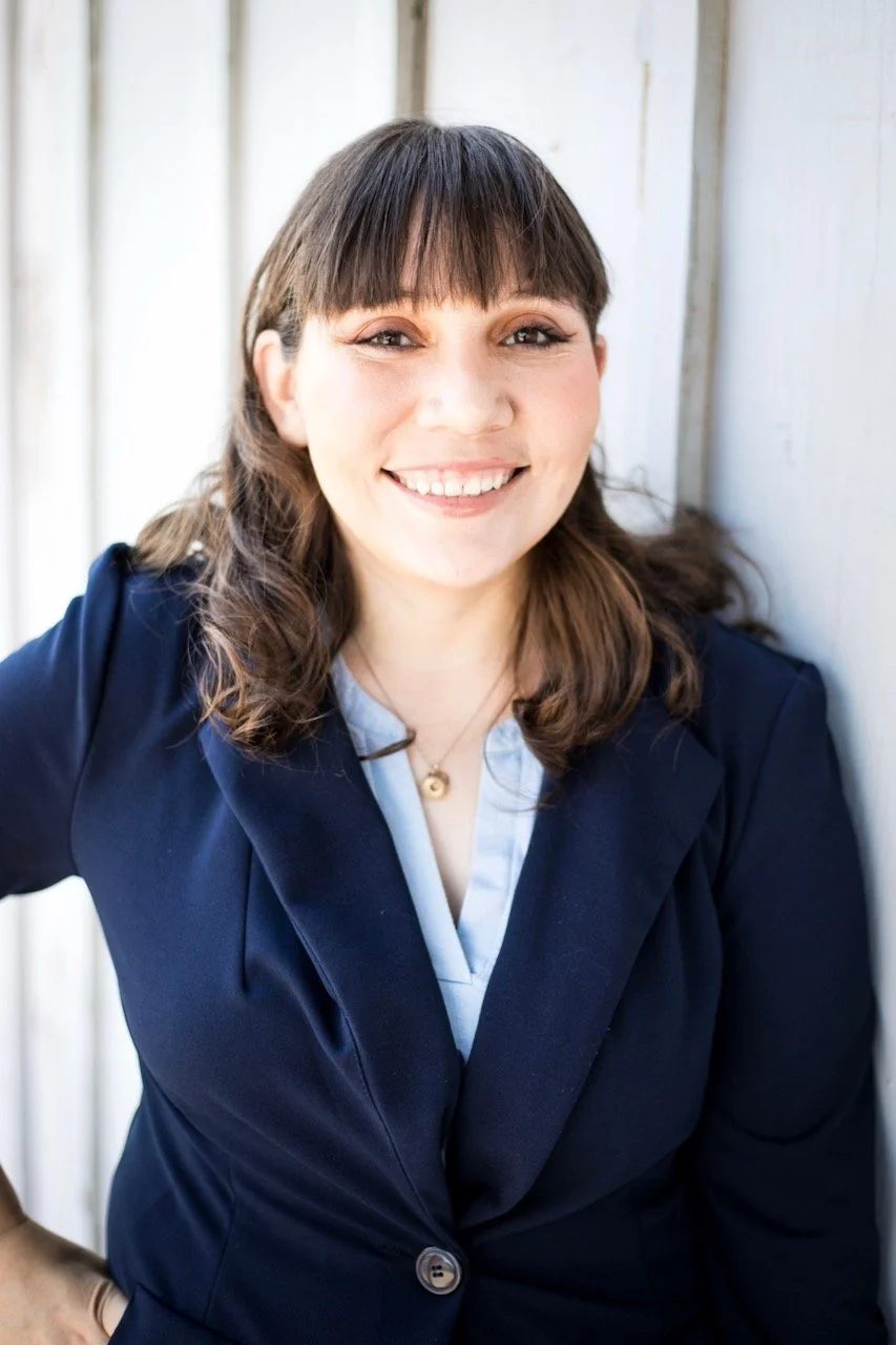 A woman with brown hair and bangs, smiling, wearing a navy blazer and white shirt, standing next to a white wood wall.