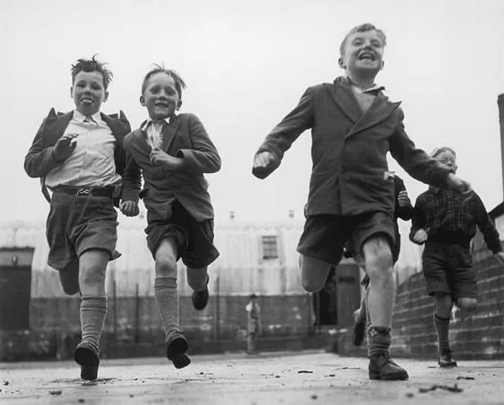 Four boys in school uniforms running and smiling outdoors, some with backpacks.