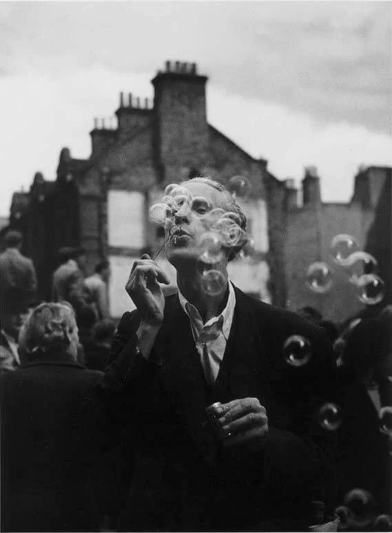 A person blowing soap bubbles on a city street with buildings in the background, black and white photograph.