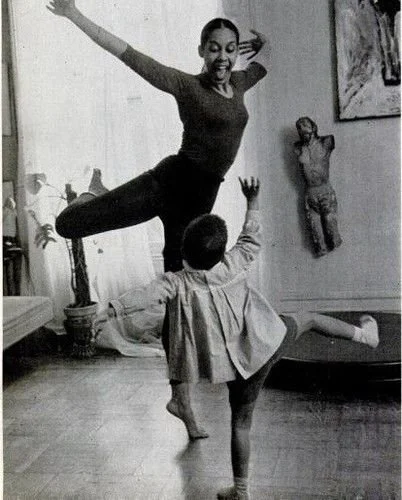 A woman is balancing on one leg with her arms extended and a big smile, while a child is in front of her with one arm raised, performing a dance or balancing move in a living room with artwork and a mannequin on the wall.