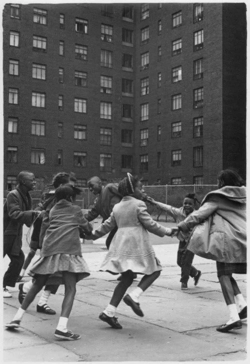 Children holding hands and playing in a circle on a playground with a tall brick building in the background.