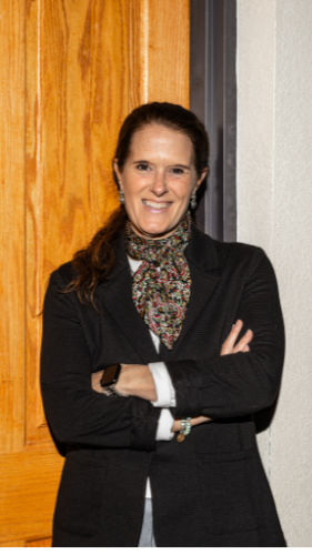 Woman in black blazer and scarf standing with arms crossed in front of a wooden door.