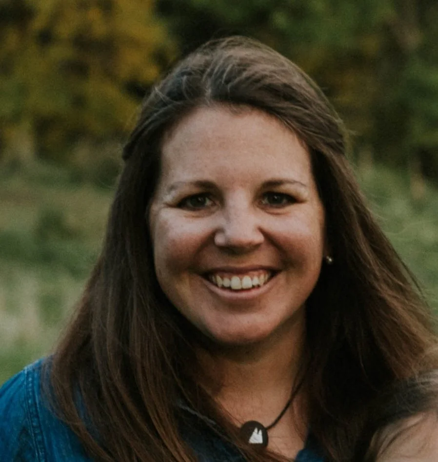 A woman with long brown hair smiling outdoors, wearing a blue top and a necklace with a mountain charm.