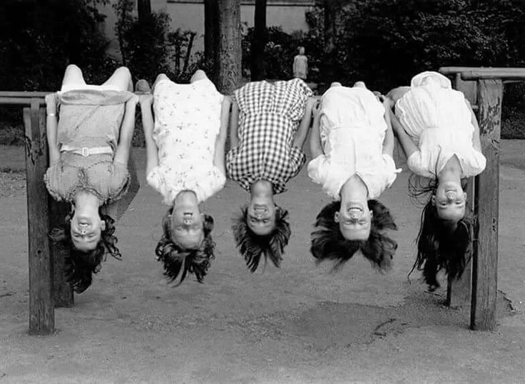 Five children hanging upside down on a horizontal bar in a park, smiling and laughing, with trees in the background.