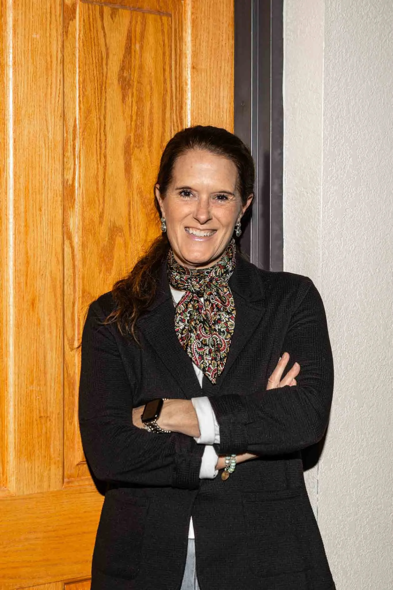 Woman in black blazer and scarf standing with arms crossed in front of a wooden door.