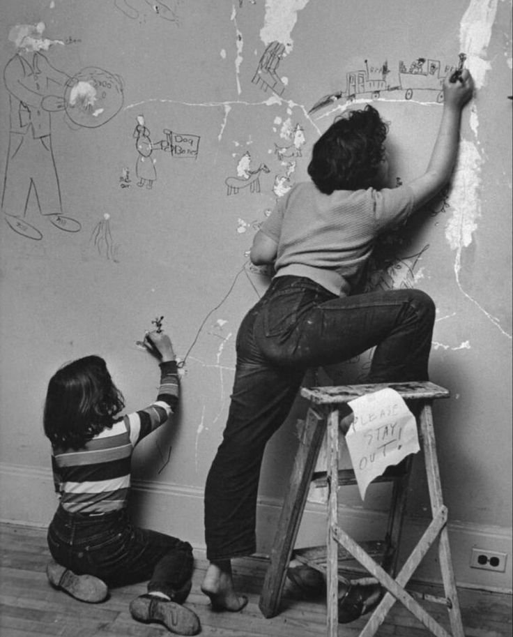 A woman and a girl drawing on a wall with chalk. The woman is standing on a small stool, reaching high to draw on the wall, and the girl is kneeling on the floor, drawing lower. The wall has various chalk sketches and a sign that says 'PLEASE STAY OUT'.