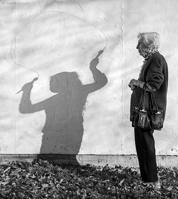 An elderly woman with gray hair, wearing a cardigan, pants, and sneakers, stands outside against a textured wall, watching the shadow of a person on the wall creating a silhouette of a woman holding a jump rope.