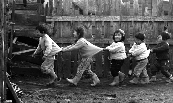 Six children holding onto each other's hips and forming a conga line, playing and smiling outdoors near a wooden fence.